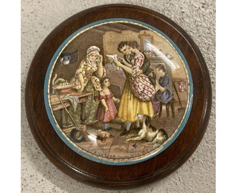 A ceramic pot lid depicting a family reading a letter in a circular wooden frame  Has a chip and crack to rear of lid. 