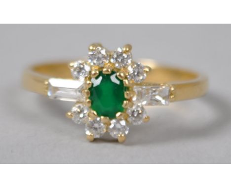 A yellow metal cluster ring, principally set with an oval cut created emerald and surrounded by circular and baguette cut col