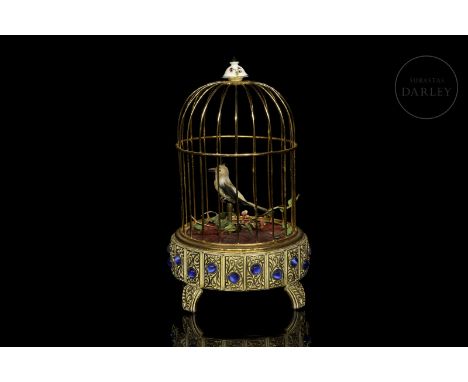 Brass cage with a small bird inside, adorned with fine feathers and arranged next to thin branches of green leaves, which has