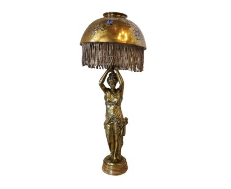 A 1920's French bronze table lamp modelled with a muse standing holding aloft the domed brass shade which is inset with facet