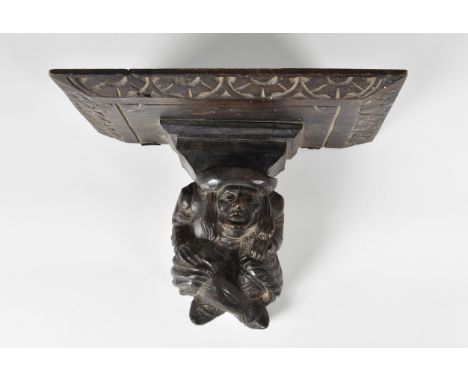 A 19th century Breton carved wooden clock bracket, in the form of a cross legged man, supporting a shelf with chamfered arcad