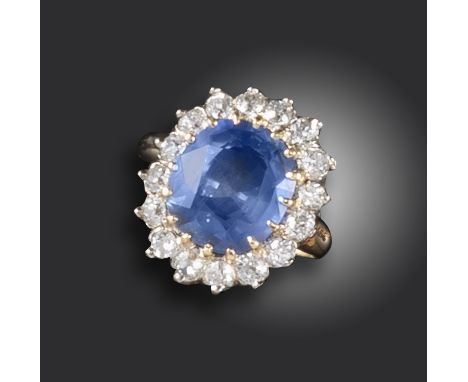 A sapphire and diamond cluster ring, the cushion-shaped sapphire is set within a surround of round brilliant-cut diamonds in 