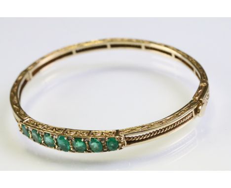 Gold emerald open hinged bangle bracelet. The bracelet being set with seven oval cut emeralds with diamond accent stones with