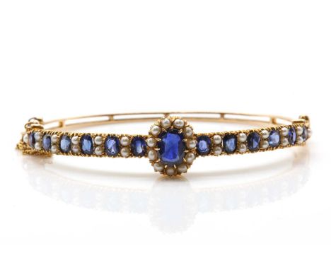 An Edwardian gold sapphire and pearl bangle,with an oval mixed cut sapphire, to a surround of pearls, all claw set. The centr