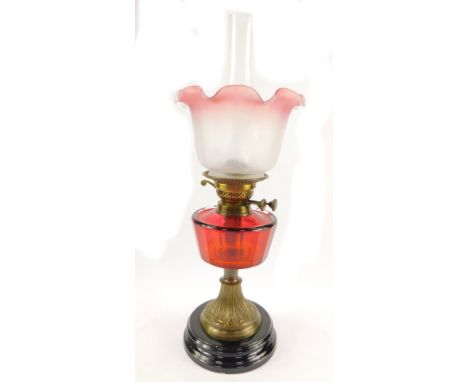 An early 20thC brass oil lamp, with a faceted ruby glass reservoir, raised on a circular black pottery socle base, with chimn