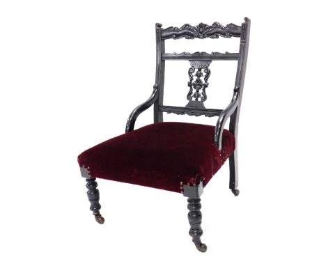 An Edwardian ebonised nursing chair, with a carved and pierced back, a red upholstered padded seat, on turned legs with ceram