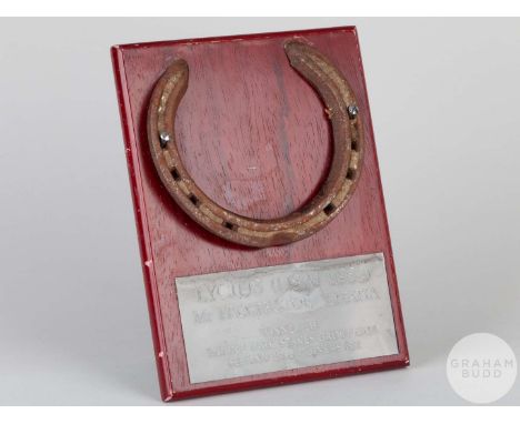 Shoe worn by the racehorse Lycius,mounted on a wooden plaque with easel support and set an engraved metal plaque, 20 by 15cm.