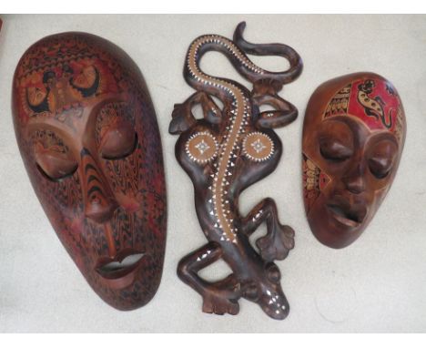 Two carved wooden wall hanging masks painted in tribal style decoration, together with a carved lizard inlaid with mother of 