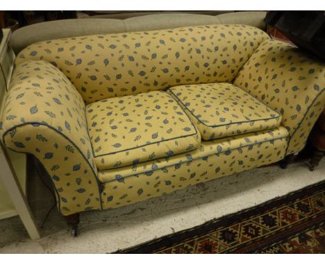 A Victorian drop-arm sofa upholstered in yellow ground and blue leaf patterned fabric, raised on turned front legs to castors