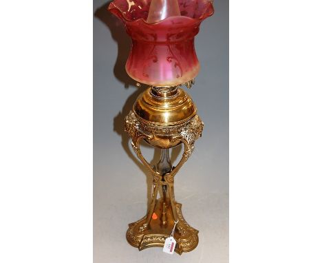 A late Victorian brass oil lamp, having cranberry tinted shade, the font with applied grotesque masks to triform base, h.67cm