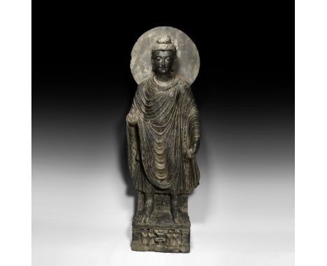 3rd-5th century AD.&nbsp;A carved schist statue of Buddha standing with loosely draped robes (sanghati) on a dais with four f