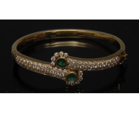 An emerald and diamond hinge bangle, cross over crest of two emerald centred terminal heads, between pave encrusted rounded b