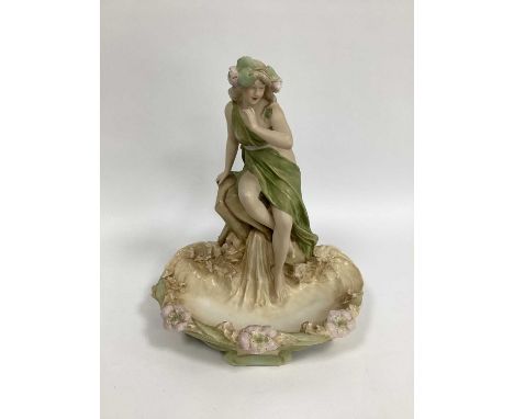 A Royal Dux Porcelain Centrepiece, modelled as a lady in a flowing dress seated on a rock beside a pond edged with flowers, p