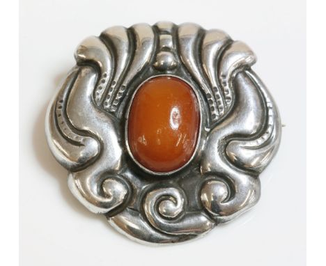 A Skonvirke Danish brooch, c.1910, with an oval cabochon amber rub set to the centre, with a repouss‚ surround of scrolls, ma