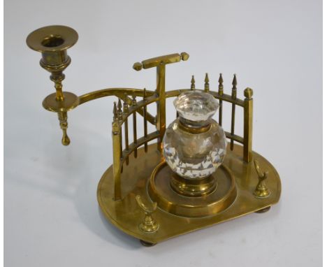 A Victorian brass ink-stand centred with a faceted glass ink bottle, with railed gallery back and hinged candle holder on tur