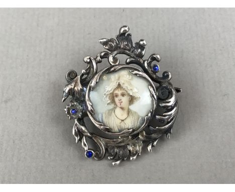 An antique white-metal brooch containing a hand-painted portrait miniature of a young lady in a lace bonnet, held in a Rococo