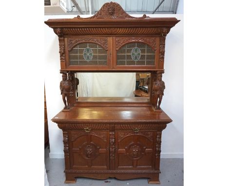 OAK MIRROR BACK SIDE CABINETwith a carved mask pediment above a pair of coloured leaded glass cupboard doors with carved scro