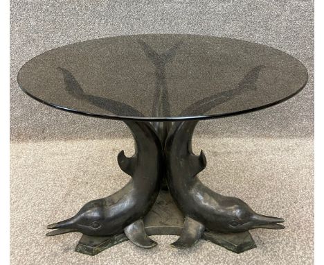 A contemporary bronze coffee table with a smoked glass circular top raised on three stylised dolphin supports. H.48 Dia.70cm.