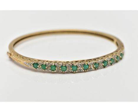 A 9CT GOLD, EMERALD AND DIAMOND HINGED BANGLE, designed with a row of eleven, claw set, circular cut emeralds, each interspac