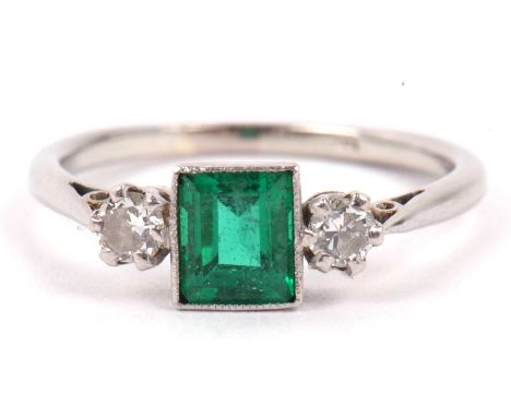 Precious metal emerald and diamond ring, the step cut emerald flanked by two small round cut diamonds, total diamond ct weigh