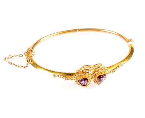 A Belle Epoque 9ct. gold bangle mounted with a pair of heart shaped amethysts within pearl borders and with a bow tied pearl 