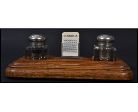A vintage early 20th century desk top pen rest and twin capstan inkwell calendar compendium. Raised on an oak base with glass