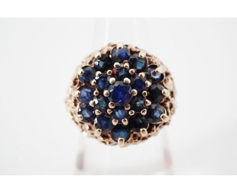 An early 1970s sapphire cocktail ring, claw-set in a tiered flower head cluster arrangement, with central sapphire of approxi