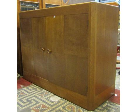 A medium to light oak side cupboard in the Heals manner, enclosed by a pair of segmented veneered panelled doors flanked by c