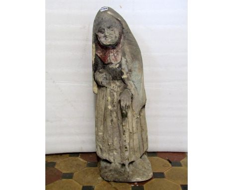 A weathered natural stone carved figure of an elderly lady wearing a cloak and pleated skirt and aided by a rustic walking st