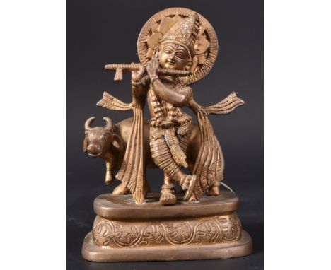 A 19th Century Indian bronze group of Krishna as a cowherd, the deity standing before a cow and playing the flute raised on e