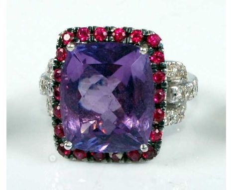 A white metal, amethyst, ruby and diamond oblong cluster ring, featuring a centre cushion cut amethyst within a border of 22 