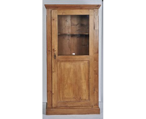A STRIPPED PINE GLAZED CUPBOARD WITH FLARED CORNICE ENCLOSED BY A SINGLE DOOR, GLAZED TO THE UPPER SECTION, PANEL BENEATH, ON