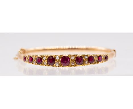 An Edwardian ruby set gold hinged bangle comprising a row of graduated grain set off round mixed cut stones, the largest appr