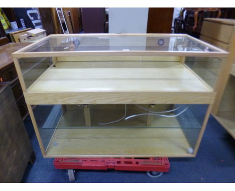 A LIGHT OAK GLAZED SHOP STYLE ILLUMINATED DISPLAY CABINET/COUNTER, the upper area with drop down access, the lower area with 