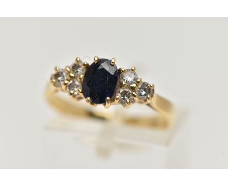 AN 18CT GOLD SAPPHIRE AND DIAMOND RING, designed with a four claw set, oval cut deep blue sapphire, flanked with three claw s