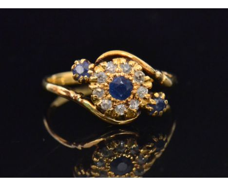 A sapphire and diamond crossover style ring with central flowerhead shaped setting of a sapphire within in border of nine old