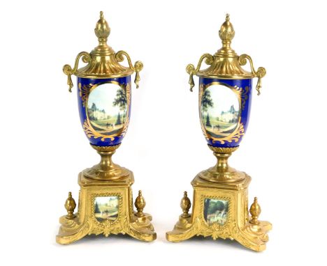 A pair of Continental urn clock garnitures, each with acorn finial the vases on a royal blue ground, depicting country houses