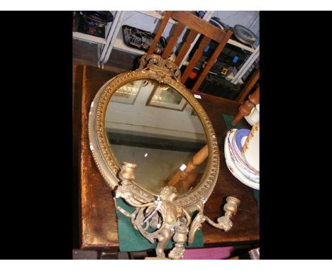 An oval mirror in ornate gilt frame sporting cherub and a three-pronged candle stick 