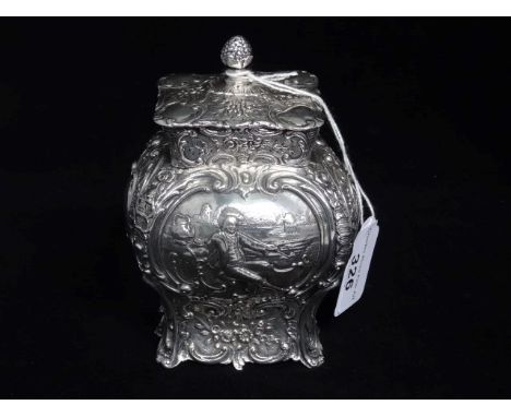 A Continental white metal tea caddy of squat baluster form, the lid with pineapple finial, the body decorated with a lady and