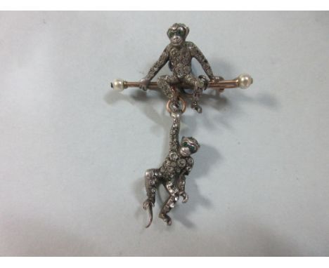 A paste set monkey brooch, the first monkey, pavé set with round white pastes and with green eyes, is sitting on a trapeze ba