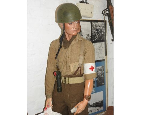 A standing mannequin wearing a shirt of plain khaki material lacking front pockets, with Medic's arm band, trouser braces, bo