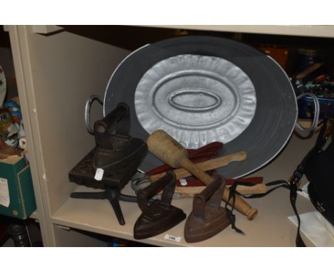 A vintage tin bath of small proportions, three cast iron flat irons , a posser, two pairs of laundry tongs and a trivet