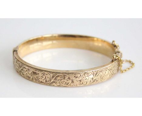 A 9ct yellow gold oval hinged hollow bangle, half engraved with scrollwork detail, with a box clasp and safety chain, bangle 