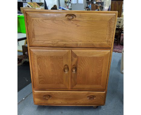 A blonde Ercol writing cabinet/bureau with a fall flap revealing a shelved interior over a pair of panelled cupboard doors wi