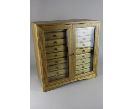 A light oak collector's tabletop cabinet of twin glazed doors enclosing two rows of nine short drawers, on a bracket base, 41