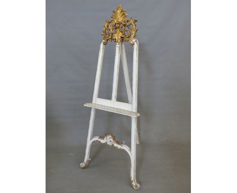 A WHITE PAINTED FLOOR STANDING  EASEL WITH GILT FOLIAGE CRESTING AND SCROLL FEET, THE PICTURE REST.   W 59cms.