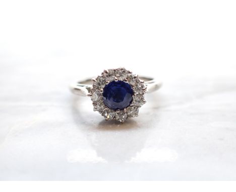 A Sapphire and Diamond Cluster Ring claw-set round sapphire within frame of eleven old-cut diamonds in white gold stamped 18c