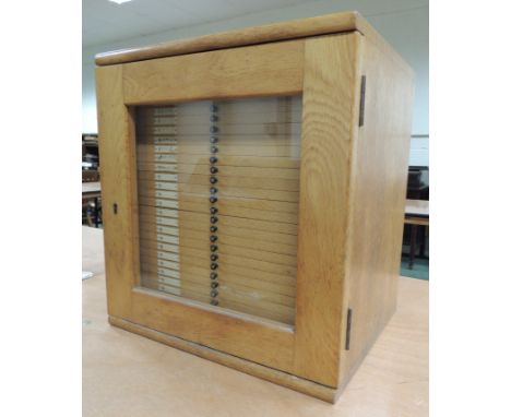 A c.1940's light oak collectors cabinet, the glazed door enclosing 28 narrow drawers, each numbered on an ivorine panel, the 