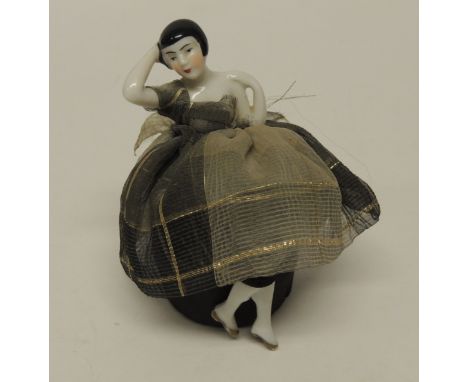 An Art Deco pin cushion half doll with legs, the glazed porcelain doll with black bob hair and in gauze dress with black cove