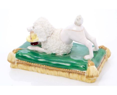 Minton poodle, circa 1830, in playful pose with basket in mouth, lying on a tasselled cushion base, 10cm long